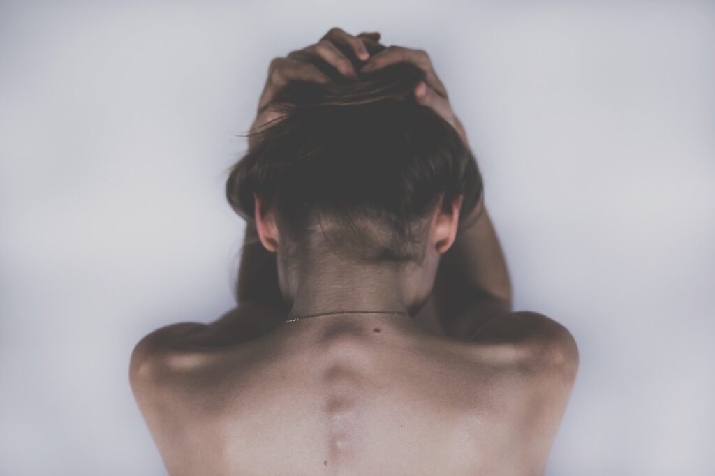 A woman with bare shoulders and visible spine structure sits with her head down and hands clasped over her head, symbolizing back or neck pain.
