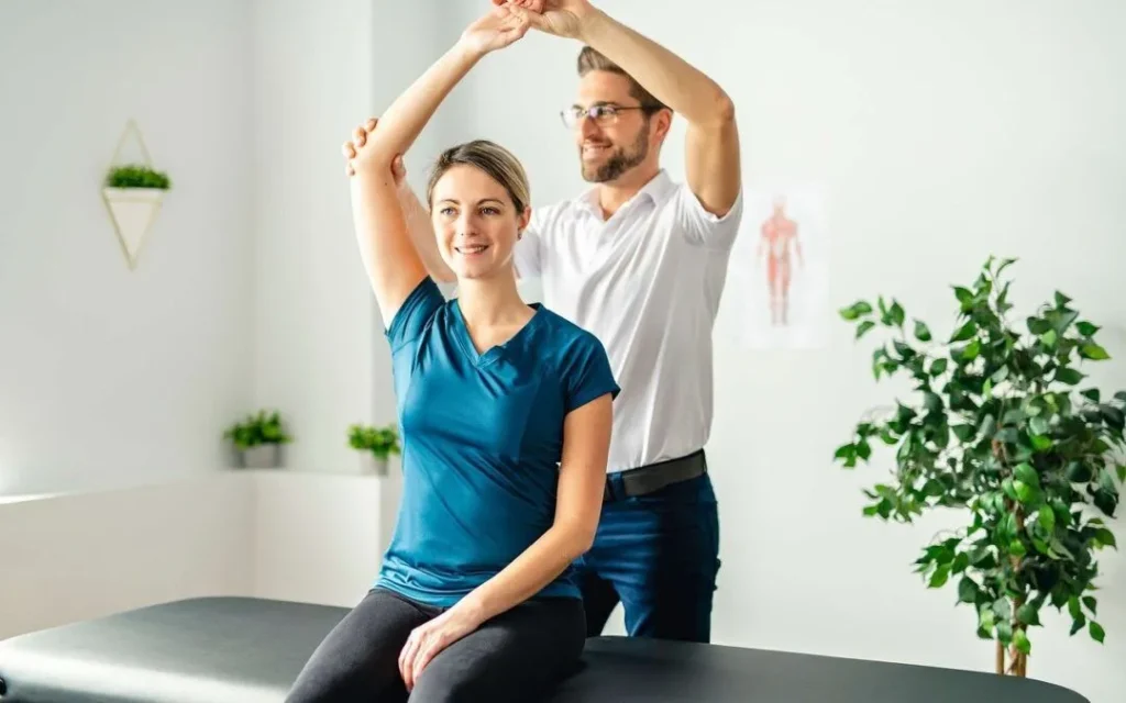 A chiropractor or osteopath helps a patient with shoulder mobility exercises in a modern clinic. The patient sits on a treatment table while the practitioner guides her arm movement.