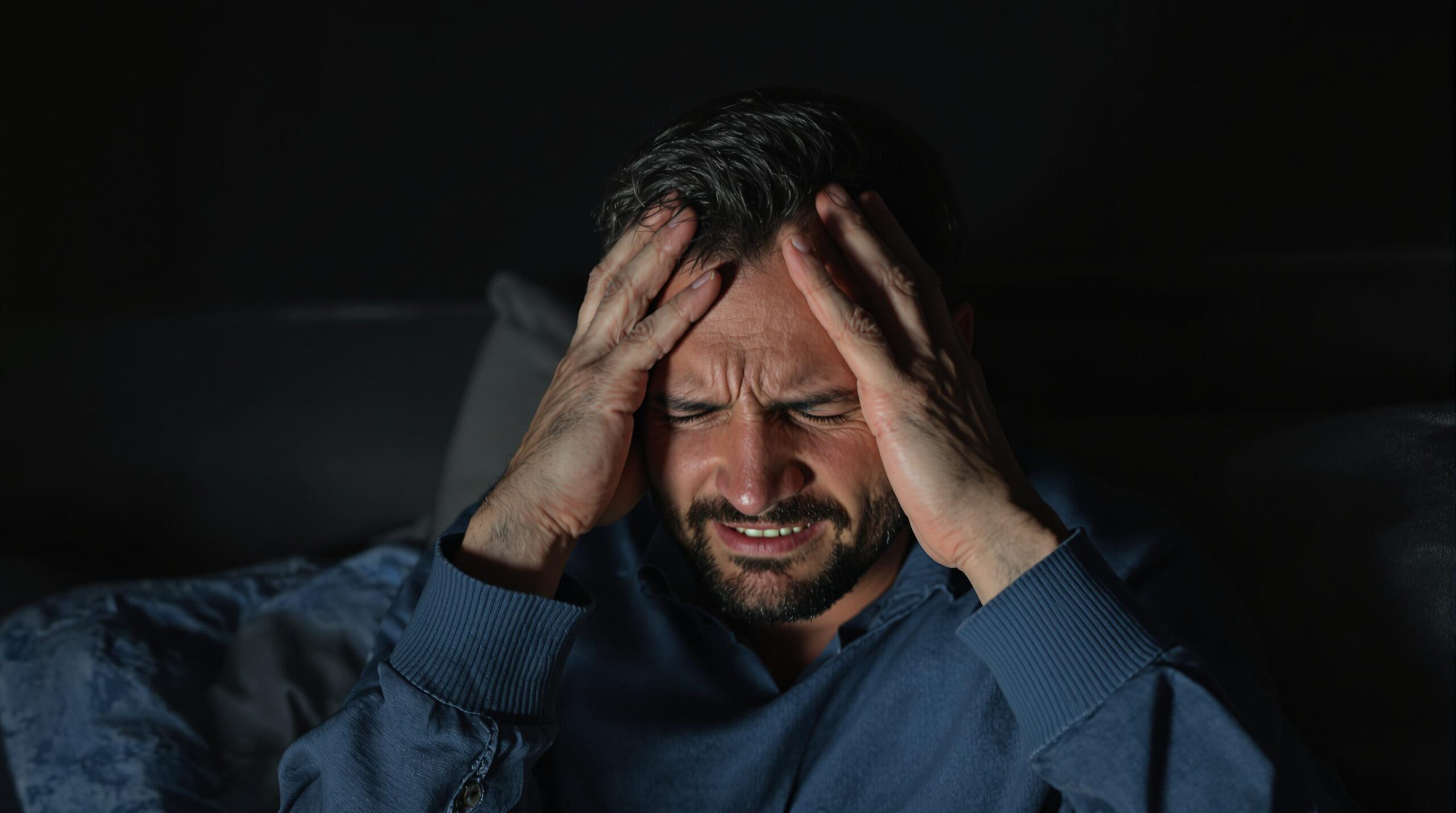 Man in a dark room holding his head in pain, showing symptoms of a severe headache or migraine.