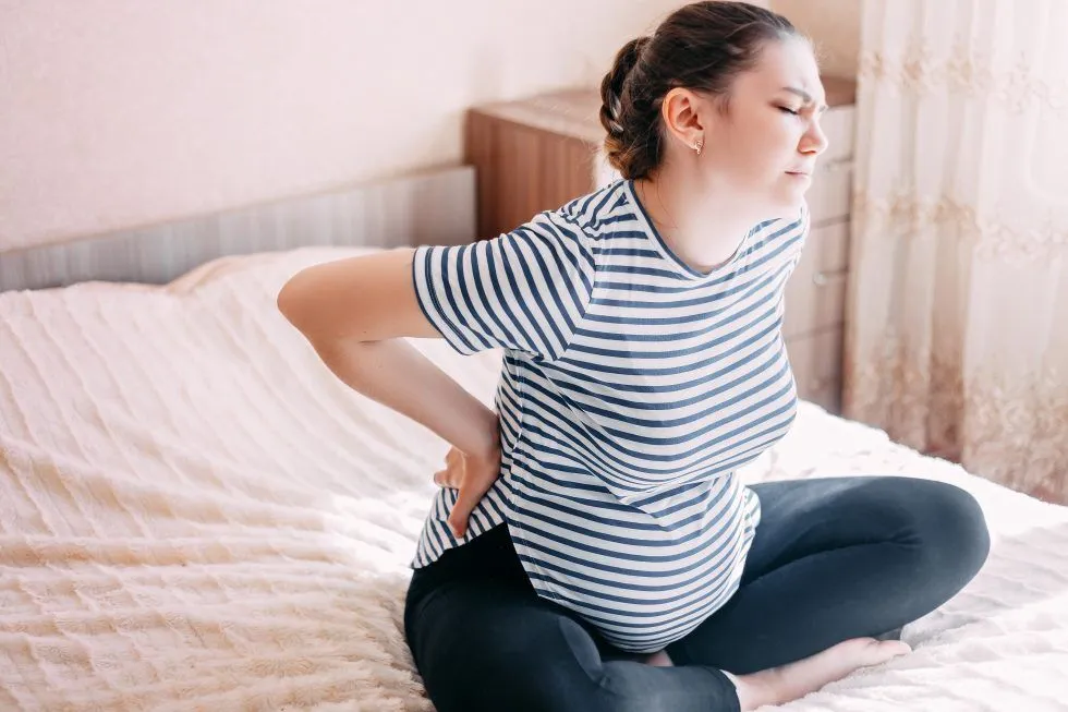Pregnant woman sitting on a bed, holding her lower back in discomfort, illustrating the need for a pregnancy chiropractor.