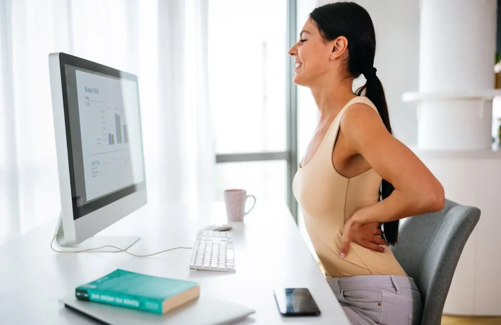 A woman experiencing back pain while sitting at her desk, holding her lower back while working on a computer.