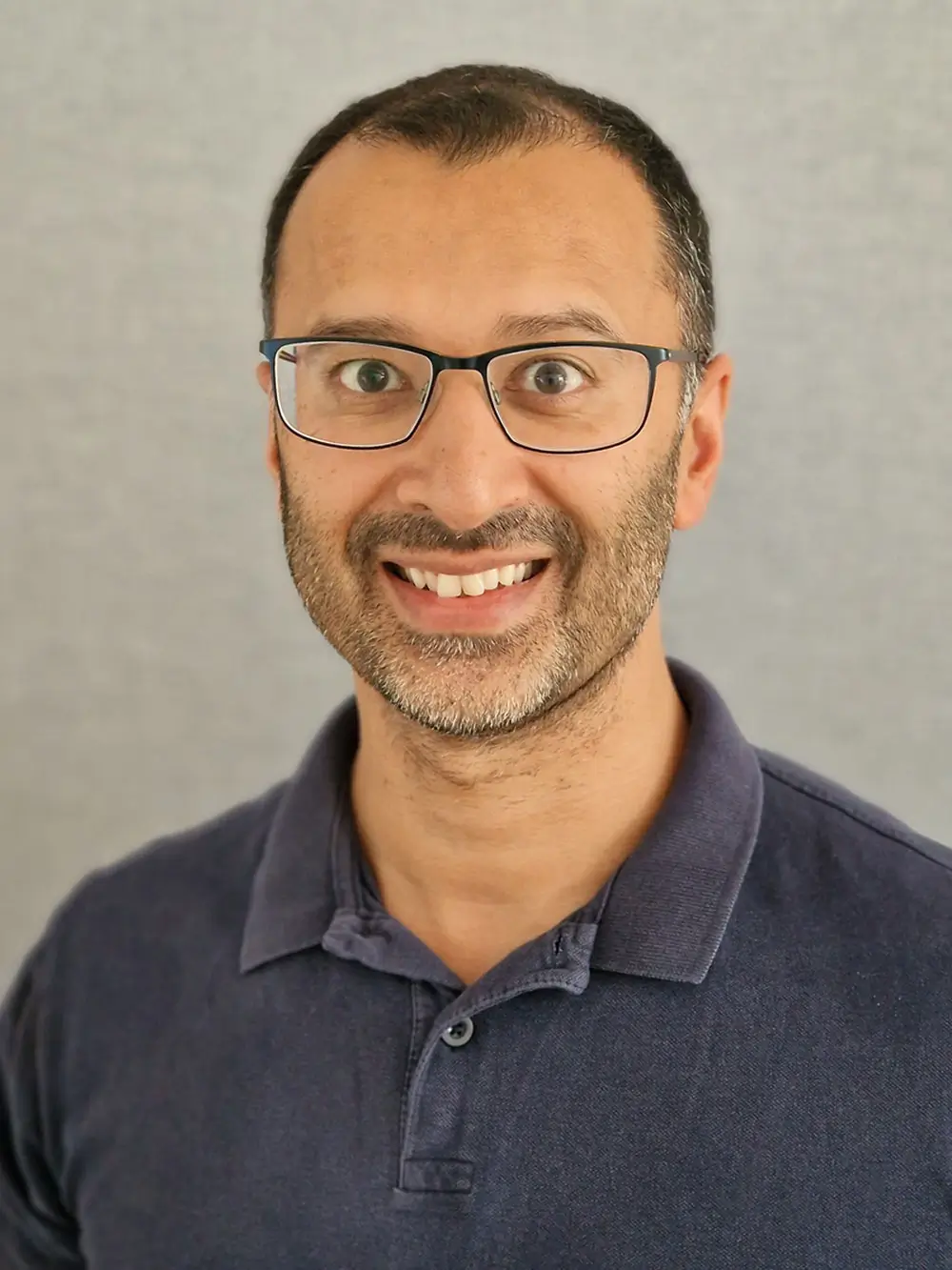 Portrait of a man with short dark hair, glasses, and a light beard, smiling at the camera while wearing a navy polo shirt against a neutral background.