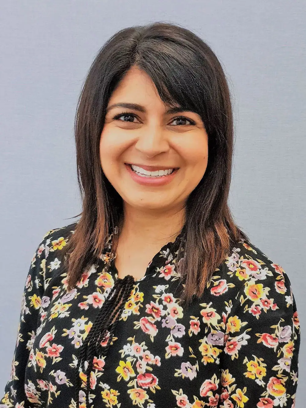 Smiling woman with shoulder-length dark hair wearing a black floral blouse, poses against a gray wall background.